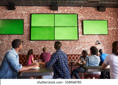 Rear View Of Friends Watching Game In Sports Bar On Screens