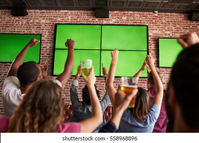 Rear View Of Friends Watching Game In Sports Bar On Screens