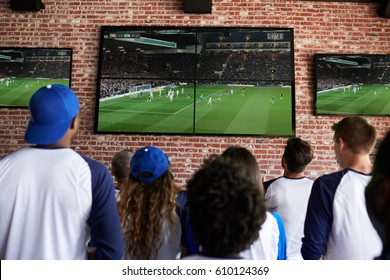 Rear View Of Friends Watching Game In Sports Bar On Screens