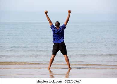 Rear View Full Body Portrait Of Excited Young Man Standing On Beach With Arms Outstretched