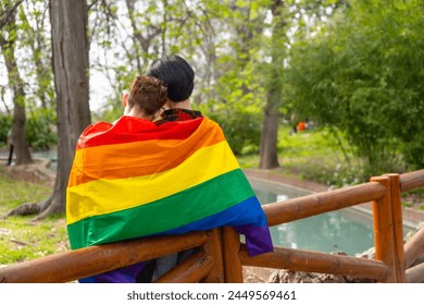 Rear view of gay couple wrapping in lgbt flag leaning on a rail a park - Powered by Shutterstock