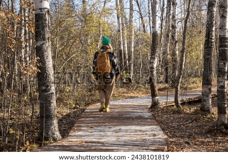 Rear view of girl with backpack walking in forest on autumn sunny warm day. Woman tourist walks along wooden road in forest and explores beauty of local nature. Student went on wildlife sanctuary