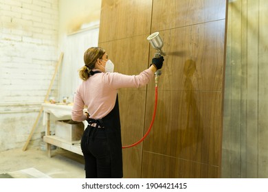 Rear View Of A Gorgeous Young Woman Holding A Spray Gun And Painting A Big Piece Of Brown Wood Panel 