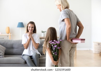 Rear View At Grandmother And Little Kid Daughter Holding Gift Box And Flowers Behind Back Congratulating Happy Mom With Mothers Day Or Birthday, Grandma And Child Making Surprise For Excited Mommy