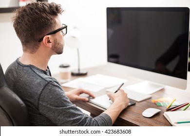 Rear View Of A Graphic Designer Using A Pen Tablet In Front Of A Computer In An Office. Blank Computer Screen