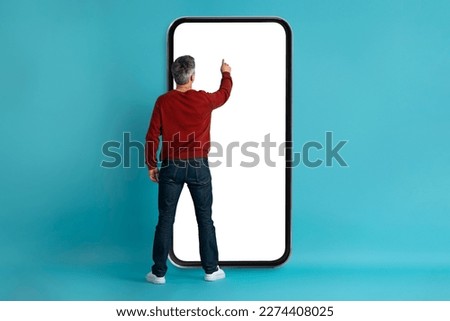 Rear view of grey-haired middle aged man in casual touching big phone white empty screen over blue studio background, checking newest mobile app, websurfing, mockup, copy space for ad