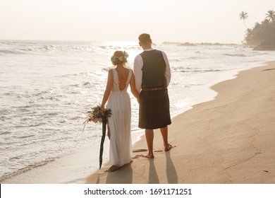 Rear View Of Groom And Bride Wearing Scottish Traditional Kilt And Bride In White Dress Walking By The Sea, Enjoy Sunset. Tropical Wedding