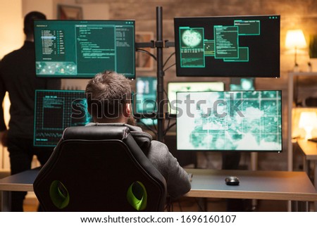 Rear view of hacker in front of computer with multiple screens in dark room.