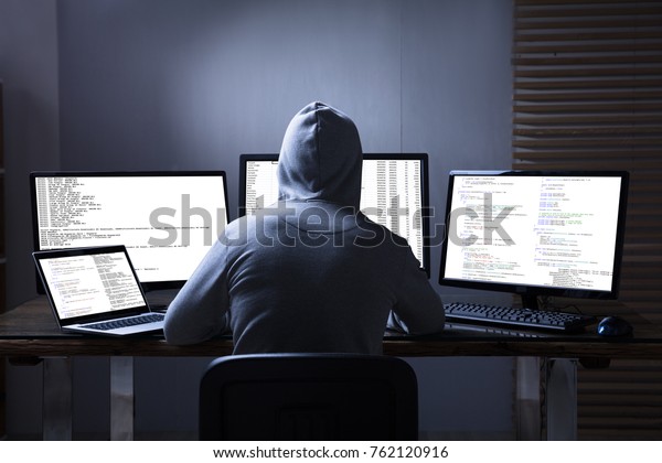 Rear View Of A Hacker Using Multiple Computers For\
Stealing Data On Desk