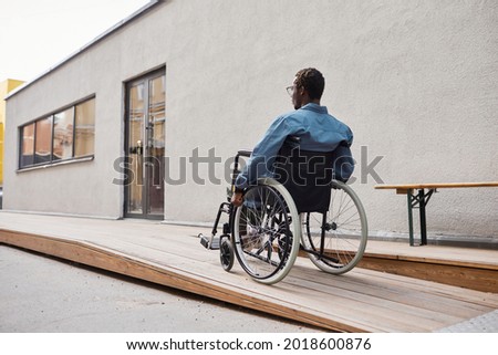 Rear view of handicapped African-American man in casual shirt using wheelchair ramp built by his house entrance