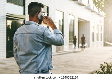 Rear View. Handsome Smiling Hipster Businessman With Beard, In Denim Jacket, Trendy Glasses Walks Around City, Calls On Mobile Phone. Happy Guy Standing Outdoor And Talking On Cell Phone. Lifestyle.