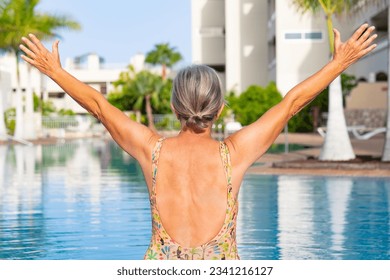Rear view of happy caucasian senior woman with outstretched arms sitting at the swimming pool enjoying freedom and vacation