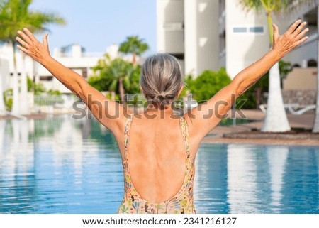 Rear view of happy caucasian senior woman with outstretched arms sitting at the swimming pool enjoying freedom and vacation