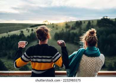 Rear view of a happy couple enjoying morning coffee and looking away while standing on a balcony with an beautiful nature in the background and with warm light at sunrise. Warm clothes, cold weather - Powered by Shutterstock