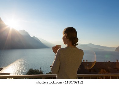 Rear view at happy free woman enjoying beautiful serene morning looking at sea lake and mountains nature landscape scenery starting new day drinking coffee relaxing. Italy, Lombardia, Riva di Solto