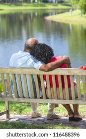 Rear view of a happy romantic senior African American couple sitting on a park bench embracing looking at a lake or river