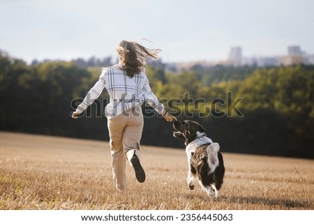Rear view of happy teen girl running with her dog across field against cityscape. Joyful pet owner with Czech Mountain Dog.