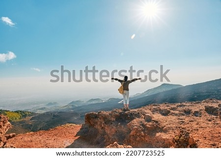 Rear view of happy tourist woman enjoying freedom with open hands, while admiring panoramic view of colorful summits of active volcano Etna, Tallest volcano in Continental Europe, Sicily, Italy.
