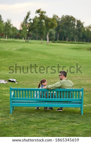 Rear view of happy young father and his cute little daughter talking while sitting together on the bench, spending time in the green park