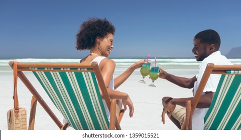Rear view of happy young multi-ethnic couple relaxing on sun lounger at beach in a sunny day. They are smiling and drinking cocktails