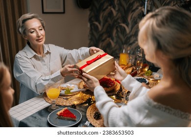 Rear view of happy young woman receiving gift from loving mother-in-law sitting with family at dinner feast table at home on Christmas Eve. Blonde female giving present to beloved daughter on holidays - Powered by Shutterstock