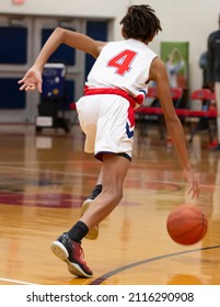 Rear View Of A High School Basketball Player Dribbling The Ball Up Court During A Game In A Gym.