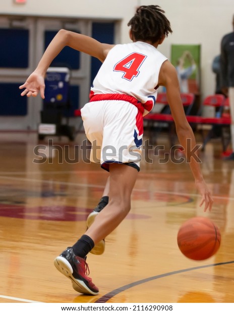 Rear view of a high school\
basketball player dribbling the ball up court during a game in a\
gym.