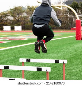 Rear View Of A High School Boy Jumping High Over Track Hurdles On A Turf Field During Strength And Agility Training In Teh Winter.