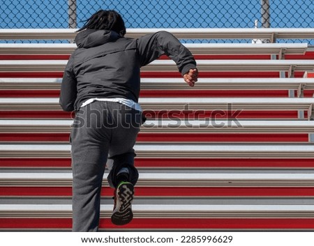 Rear view of a high school boy running up bleachers wearing long black pants and long sleeves during track practice in the early spring in New York.