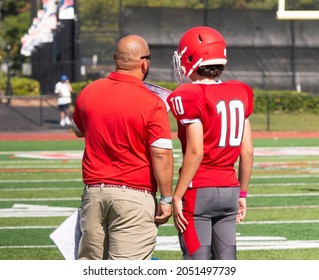 Rear view of a high school head football coach going over the next play with his quarterback on the sidline during a game.