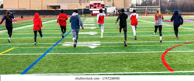 Rear View Of High School Track Team Of Boys And Girls Performing Warm Up Running Drils Side By Side On A Greeen Turf Field.