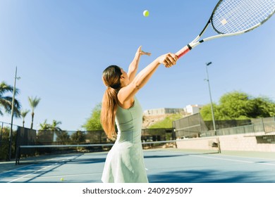 Rear view of an hispanic young woman practicing a good tennis serve with a racket while exercising on the tennis court - Powered by Shutterstock