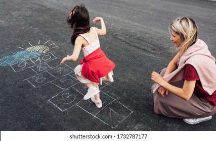 Rear View Image Of Little Girl Playing Hopscotch With Her Mother On Playground Outdoors. Child Plays With Her Mom Outside. Kid And Woman Plays Hopscotch Drawn On Pavement. Activities And Games Outside