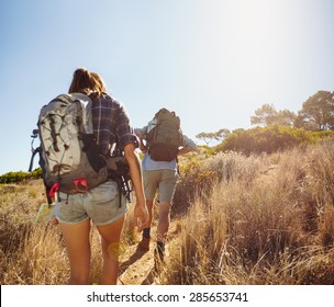 Rear View Image Of Two Young People Carrying Backpacks Walking Through Mountain Trial On Summer Day. Man And Woman Hiking On Bright Sunny Day.