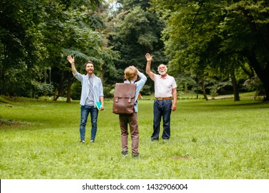 Rear View Of Kid With Backpack Going To School And Waving To Parents - Father And Grandfather Saying Goodbye Their School Boy. Back To School Concept. First Day Of Fall.