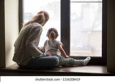 Rear View At Kid Daughter And Single Mother Sitting On Sill Dreaming Of Good Future Concept, Babysitter, Nanny Or Young Mom And Child Girl Looking Outside Window Thinking Of New Goals Feeling Hopeful