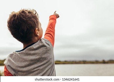 Rear View Of A Kid Standing Near The Lake And Showing Something To Someone. Close Up Back View Of A Boy Pointing His Finger To The Sky.