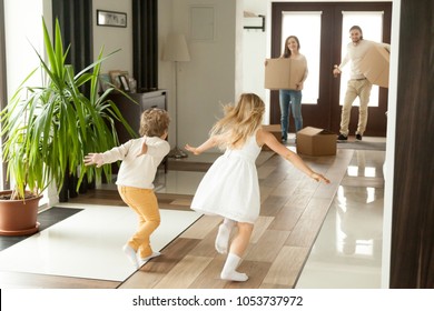 Rear View At Kids Running To Parents Holding Boxes On Moving Day Or After Shopping, Excited Happy Children Son And Daughter Having Fun In New Home Enjoying Relocation, Exploring Luxury House Together