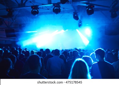 Rear View Of Large Crowd Of Men And Women Standing Motionless In Dark Hall In Blue Light Before Stage, Back Lit