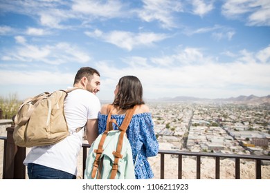 Rear View Of Latin Backpackers Couple Looking At City From Height And Lost In Deep Conversation