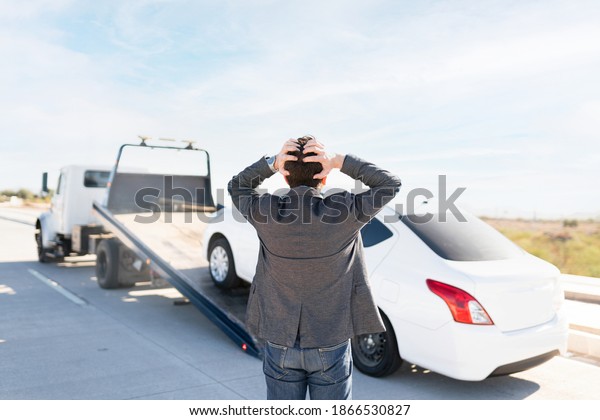 Rear view of a latin man in his 30s looking angry\
because he is watching his car being towed on a tow truck from the\
side of the highway