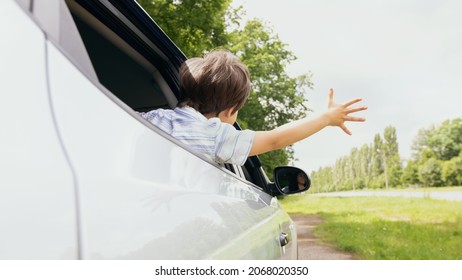 Rear View Of Little Boy Leaning Out Of Car Window And Trying To Catch Wind With Outstretched Hand, Small Male Child Enjoying Summer Weather And Automobile Ride, On The Move Shot, Closeup