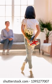 Rear View Little Daughter Holding Flowers, Bouquet And Gift Behind Back, Congratulate Young Mother With Mothers Day Or Birthday, Adorable Child Making Surprise To Mum At Home, Vertical Photo