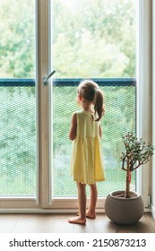 Rear view of a little girl in yellow dress standing near the window and looking through it - Shutterstock ID 2150873213
