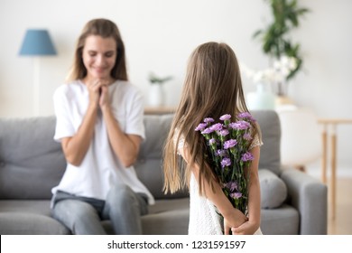 Rear View At Little Kid Daughter Holding Violet Flowers Congratulating Happy Young Mom With Mothers Day Or Birthday, Child Girl Hiding Bouquet Behind Back Making Surprise To Grateful Mommy At Home