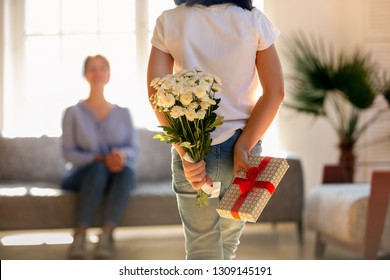 Rear View At Little Kid Daughter Holding Flowers And Gift Box Behind Back Congratulating Happy Young Mom With Mothers Day Or Birthday, Child Girl Hiding Bouquet Present Making Surprise To Mum At Home