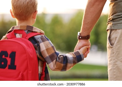 Rear view of little son schoolboy with backpack holding hand of father dad while going to first grade on sunny autumn day, cropped shot of parent and small child on the way to school, standing in park