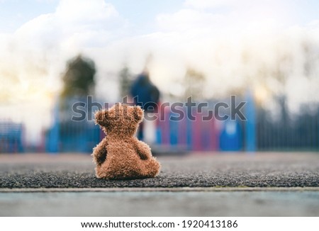 Rear view Lonely Teddy bear doll sitting alone on footpath with blurry kid playing playground in retro filter, Back view Lost brown bear toy looking at people, International missing children's day