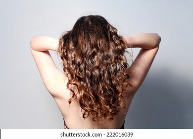 Rear View Of The Long Curly Haired Women In Front Of Light Gray Wall With Her Hands Combing Her Hair. Back Side Photo Of Young Woman With Long Curly Brown Hair Semi-nude