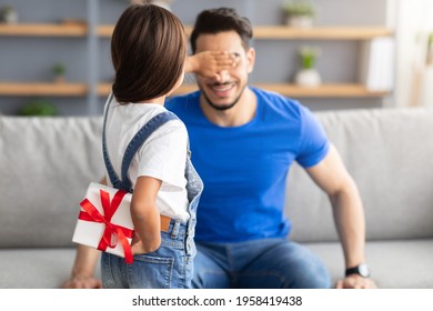 Rear view loving daughter holding and hiding wrapped gift box behind her back, making surprise for excited dad, covering his eyes, greeting young man with father's day or birthday, guy sitting on sofa